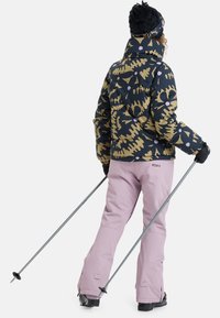 Gevoerde skijas in marineblauw met beige en roze abstracte patronen; gecombineerd met lichtroze skibroeken en zwarte handschoenen. Skistokken in de hand.