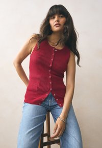 Red ribbed sleeveless top with a button front, featuring a smooth texture and contrasting silver buttons, paired with light blue jeans.