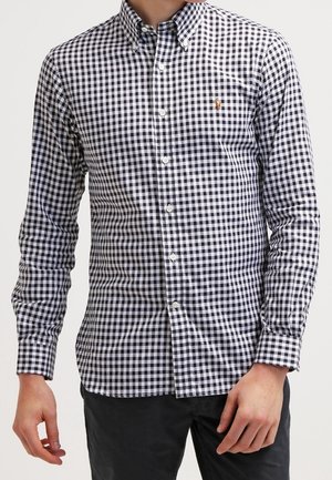 Uomo che indossa una camicia a maniche lunghe con bottoni a quadri bianchi e neri e pantaloni scuri, in piedi con una mano rilassata.