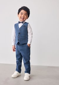 Conjunto formal de niño que incluye un chaleco azul con botones, camisa blanca, pajarita oscura y pantalones a juego. Completado con zapatillas blancas.