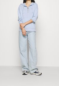 Sudadera con capucha de color azul claro, con cremallera frontal, mangas largas y un bolsillo tipo canguro. Combinada con pantalones vaqueros de pierna ancha de color azul claro y zapatillas.