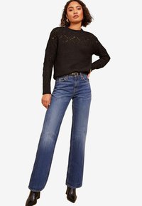 Schwarzer Strickpullover mit dekorativem Muster, kombiniert mit hochgeschnittenen blauen Jeans im Schlagstil und schwarzen stiefelartigen Absatzboots.
