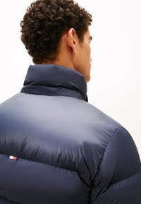 Marineblauer Daunenjacke mit hohem Kragen, die eine glatte, glänzende Textur aufweist und ein kleines Logo in Rot, Weiß und Blau auf der Rückseite hat.