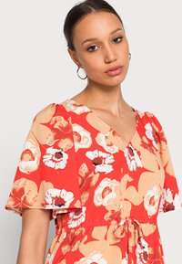 Vestido floral vermelho com mangas curtas, apresentando um decote em V e uma cintura atada. O tecido tem uma textura suave com flores grandes em branco e bege.