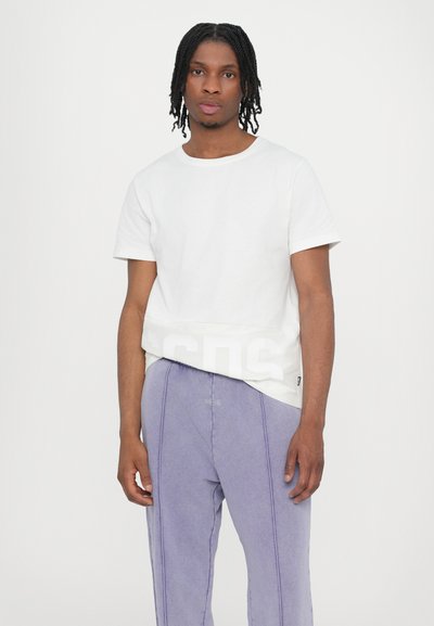 White short-sleeve t-shirt with a rounded neckline and white lettering on the hem, paired with light purple pants featuring contrasting seams.