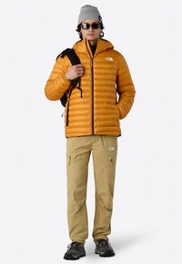 Giacca isolante gialla con zip frontale, dettagli neri, indossata sopra una maglia nera a maniche lunghe. Pantaloni beige con tasche e calzature nere.