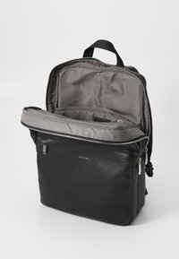 Mochila de cuero negra con interior de tela gris, apertura con cremallera y diseño minimalista. Presenta un logo y un asa superior para llevar.