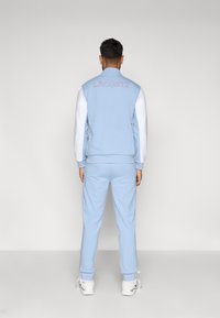 Γαλάζιο αθλητικό σύνολο φτιαγμένο από βαμβάκι με λευκά μανίκια. Διαθέτει κεντημένο λογότυπο "LACOSTE" στην πλάτη και ριγέ λεπτομέρειες παντελονιού.