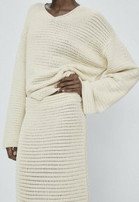 Personne à la peau foncée portant un pull et une jupe crochetés de couleur crème, une main remontant légèrement le pull.