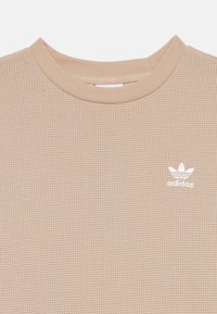 Beżowy t-shirt z krótkim rękawem wykonany z teksturowanej tkaniny w splot wafelowy. Posiada białe haftowane logo na lewej piersi oraz ribbowany dekolt.