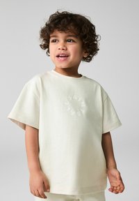 Lys beige bomulds t-shirt med løs pasform, der har et cirkulært grafik, som står "SAINT" i en diskret tone. Korte ærmer og blød tekstur.