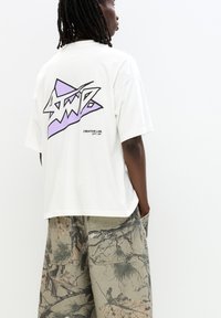 T-shirt bianco oversize con un design grafico viola e nero sulla schiena. Abbinato a pantaloni cargo verdi con motivo.