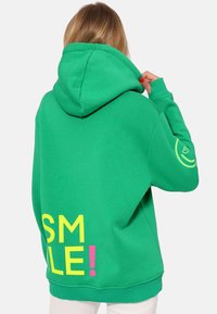 Felpa con cappuccio verde realizzata in cotone. Presenta grafiche audaci in giallo e rosa con la scritta "SORRIDI!" sul retro e un logo circolare sulla manica.