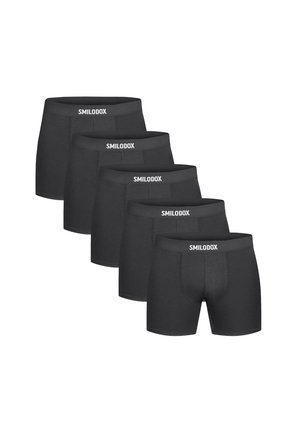 Muskelbepackter Mann trägt schwarze Boxershorts und weiße Knöchelsocken, steht vor einem einfachen hellen Hintergrund und schaut zur Seite.
