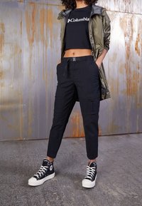 Svarta cargobyxor med flera fickor, kombinerat med en kortärmad svart t-shirt med en vit logotyp och svarta hög-top sneakers. Olivergrön jacka.