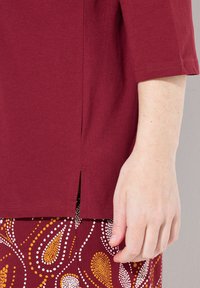 Bordeaux katoenen top met driekwartmouwen, voorzien van een zijsplit. Hand zichtbaar, met een lichte huidskleur, tegen een geblokte rok.