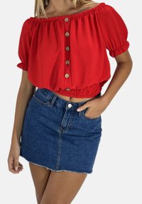 Haut rouge court, décolleté épaules nues, avec manches courtes bouffantes et boutons en bois ; assorti à une mini-jupe en denim cintrée avec un ourlet effiloché.