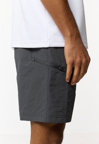 Άνδρας που φοράει σκούρα γκρι σορτς με πλαϊνή τσέπη με φερμουάρ και λευκό πουκάμισο, στέκεται μπροστά σε απλό φόντο.