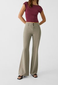 Vrouw in beige geruite hoge taille flare broeken, maroon cropped aansluitend topje en zwarte open teen hakken, staand met handen achter haar rug.