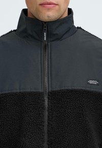 Giacca nera con zip, caratterizzata da una parte inferiore morbida e testurizzata e una parte superiore liscia. Collo alto e un piccolo logo sul petto.