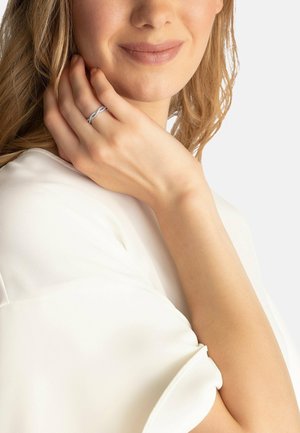 Silberner Ring mit einem verzwirbelten Design, getragen an einer Hand mit heller Haut. Der Hintergrund hat eine neutrale Farbe und zeigt einen weißen, glatten Stoff.