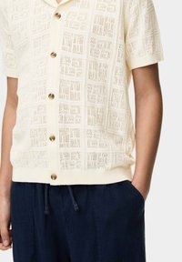 Camisa de punto de manga corta en color crema con un patrón en forma de encaje y cuello redondeado, que cuenta con cuatro botones marrones y dobladillo acanalado, llevada sobre pantalones azul marino.