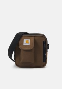 Brązowa torebka typu crossbody z płótnа, z zamkiem błyskawicznym, przednią kieszenią z klapą i regulowanym czarnym paskiem. Zawiera naszywkę z logo Carhartt.