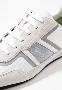 Vit och grå sneaker med mocka, mesh och syntetmaterial, med platta vita skosnören och en texturerad grå sidopanel.