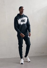 Mann iført svart Nike-hettegenser, matchende joggebukser og hvite Nike-sneakers står mot en enkel lysvegg.