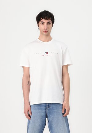 T-shirt en coton blanc avec un col rond et des manches courtes. Présente un logo qui comprend un motif rectangulaire rouge et bleu. Jeans bleus.