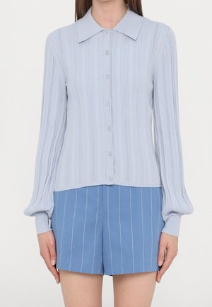 Vrouw draagt een lichtblauwe cardigan met knopen en verticale patronen en blauwe shorts met fijne strepen tegen een witte achtergrond.