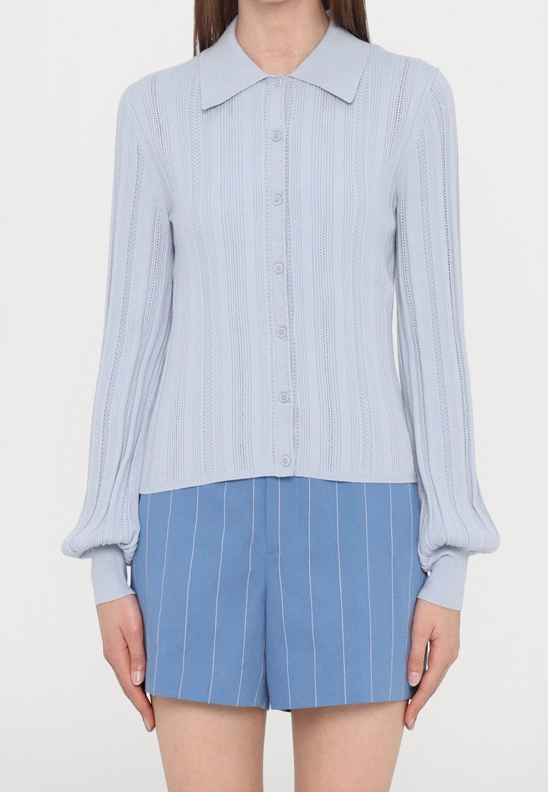Femme portant un gilet bleu clair boutonné avec des motifs verticaux et un short bleu à fines rayures sur fond blanc.