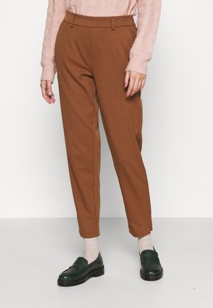 Kobieta w brązowych spodniach o wysokim stanowią, zwężanych ku dołowi, jasnoróżowym swetrze z warkoczowym splotem, beżowych skarpetkach i ciemnozielonych mokasynach, stojąca na szarej podłodze.