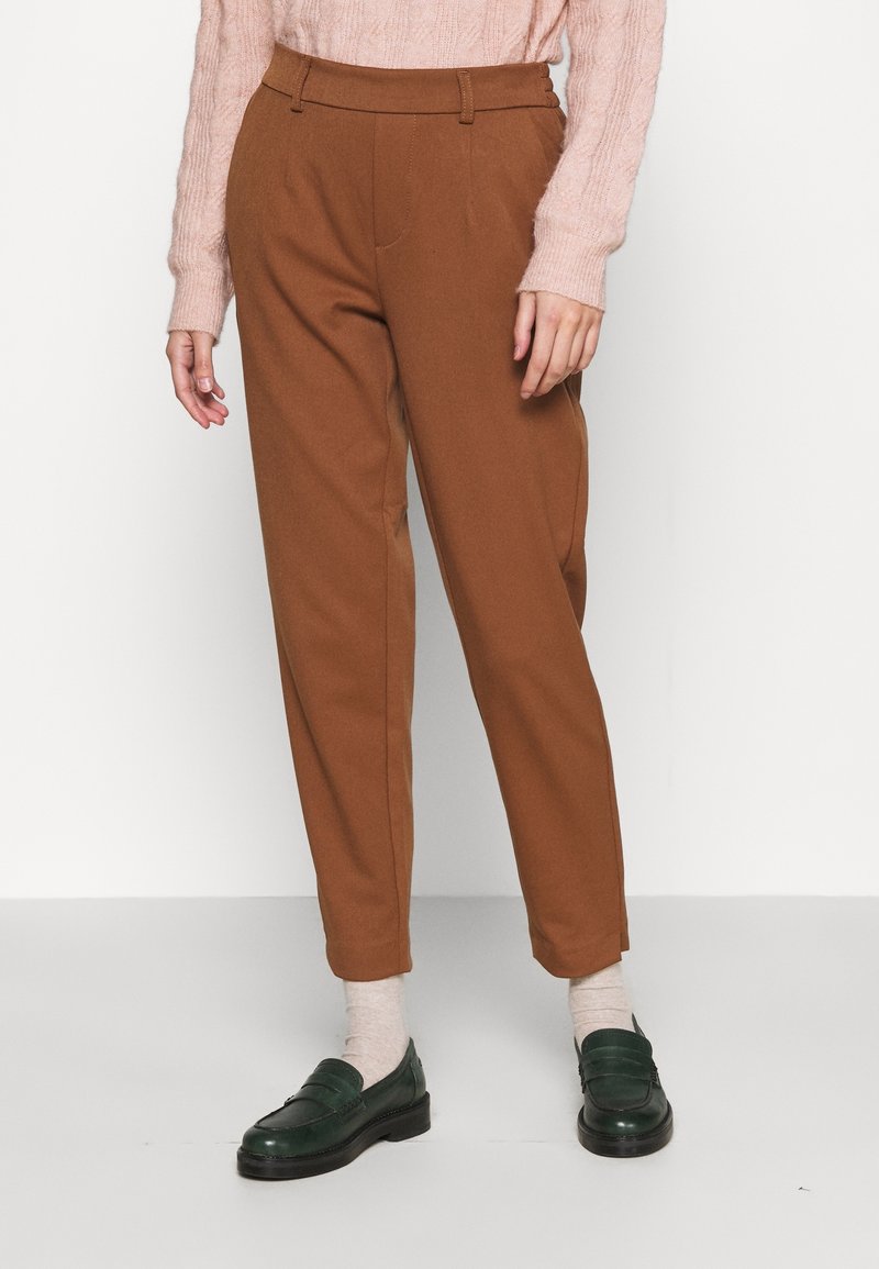Femme portant un pantalon marron taille haute à coupe effilée, un pull en maille torsadée rose clair, des chaussettes beiges et des mocassins vert foncé, debout sur un sol gris.