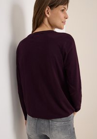 Lila Langarmshirt mit einem runden Halsausschnitt, mit einem lockeren Schnitt. Kombiniert mit hellgrauen Jeans, von hinten vor einem neutralen Hintergrund zu sehen.