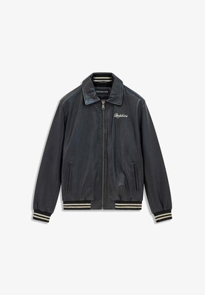 Veste aviateur en cuir noir avec fermeture éclair, poignets et ourlet côtelés ornés de rayures blanches, et le mot "Redskins" brodé sur la poitrine.