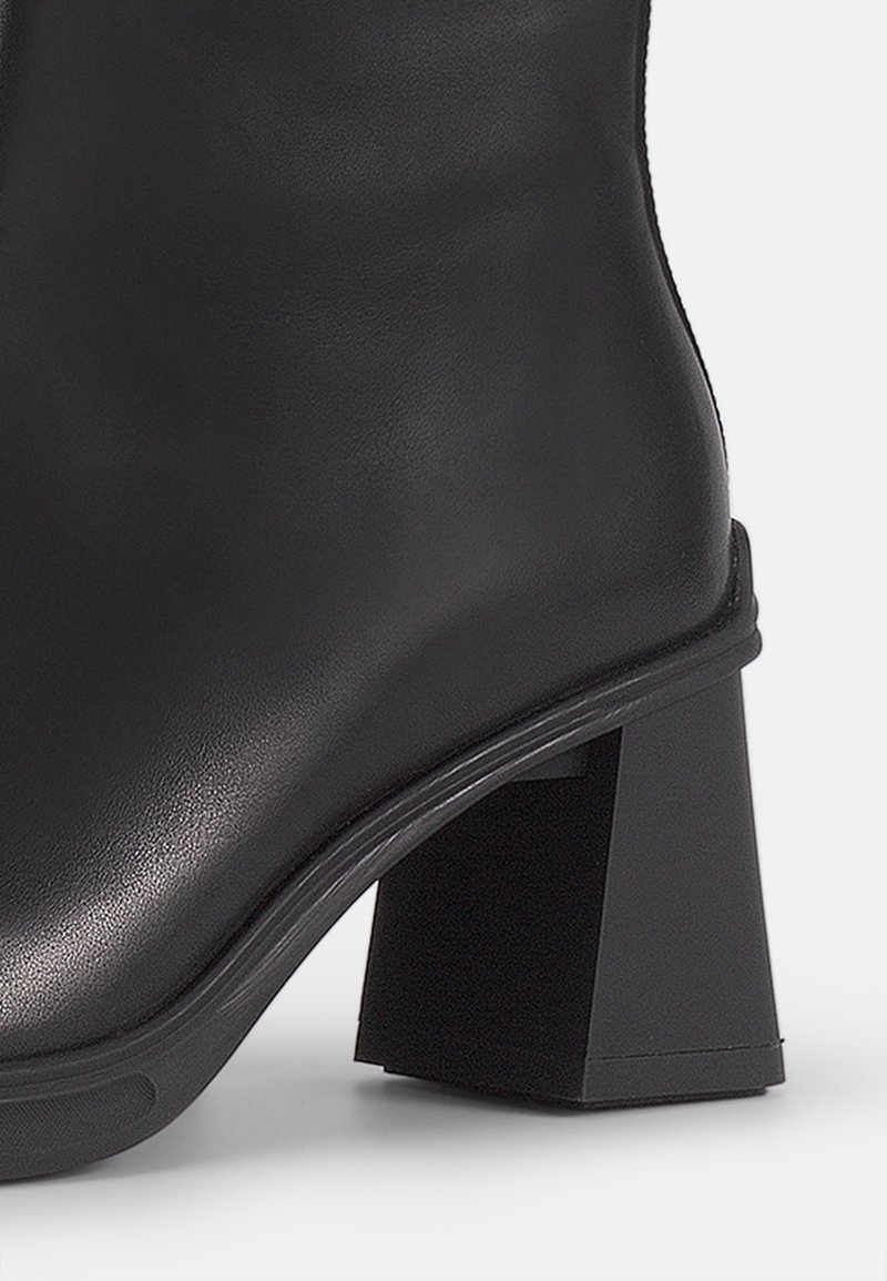 Botín negro de tobillo hecho de cuero suave con un tacón cuadrado, que presenta un brillo sutil y costuras mínimas para un diseño elegante.