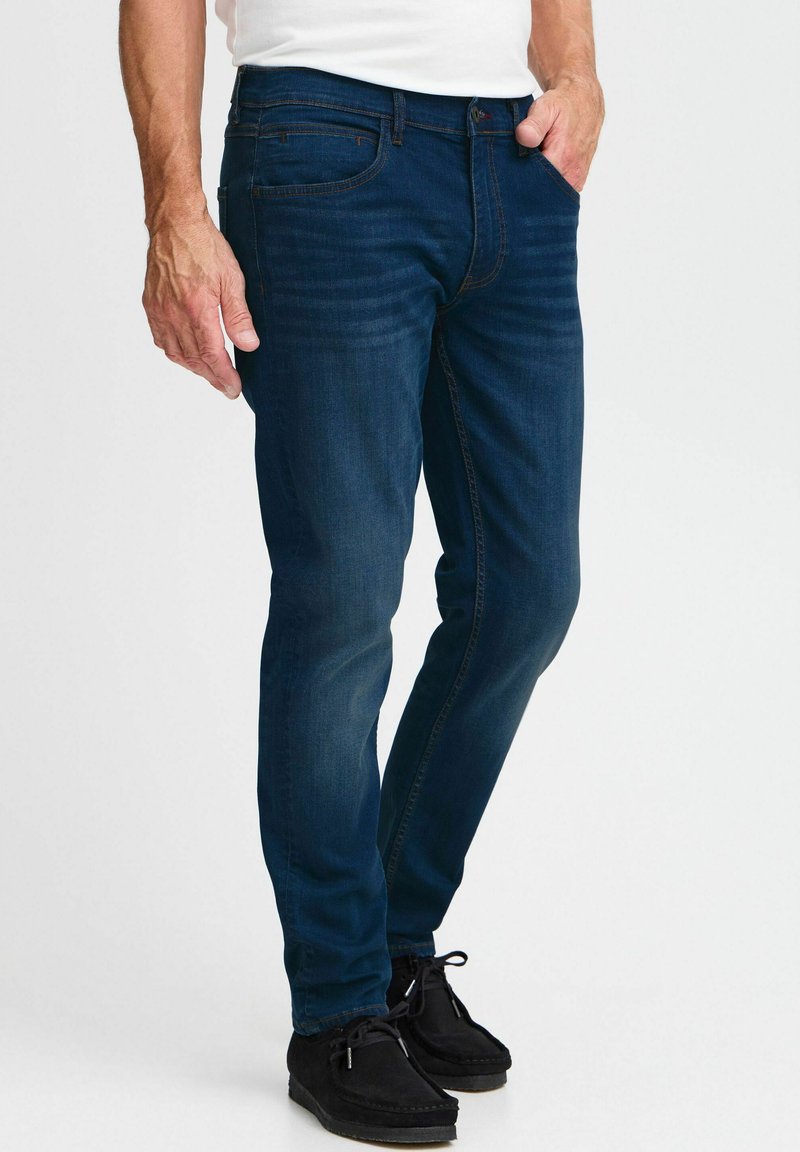 Jeans de mezclilla azul oscuro con corte ajustado, que cuentan con cinco bolsillos, un ligero desgaste en los muslos y costuras en contraste; combinados con zapatos negros.