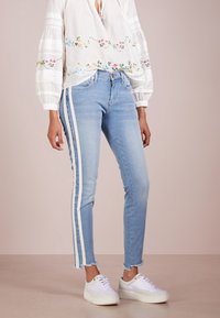 Jeans de mezclilla azul claro con franjas blancas laterales, con un dobladillo ligeramente deshilachado y un diseño ajustado. Combinados con zapatillas blancas.