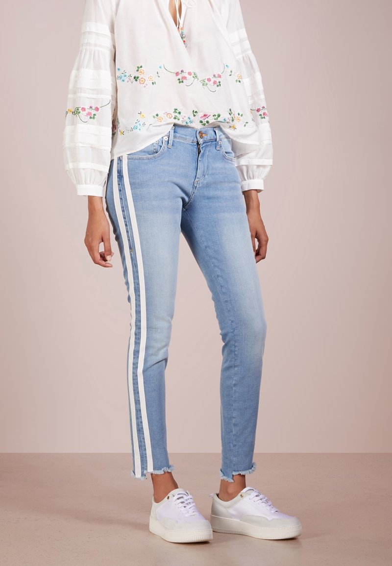 Jeans de mezclilla azul claro con franjas blancas laterales, con un dobladillo ligeramente deshilachado y un diseño ajustado. Combinados con zapatillas blancas.