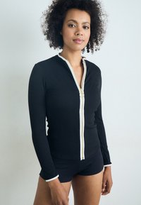 Black ribbed long-sleeve top with a zip front, white accents, and round zipper pull. Paired with black shorts, displaying a fitted design.
