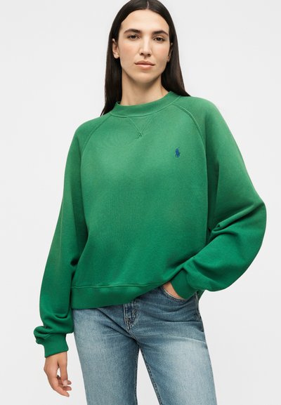 Γυναίκα με μακριά σκούρα μαλλιά, ντυμένη με πράσινο φούτερ με στρογγυλή λαιμόκοψη και ανοιχτό μπλε τζιν, στέκεται με το ένα χέρι στην τσέπη, μπροστά από απλό φόντο.