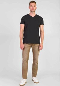 Zwarte v-hals t-shirt, lichtbruine jeans, witte sneakers met bruine zolen; model staat rechtop tegen een effen grijze achtergrond.