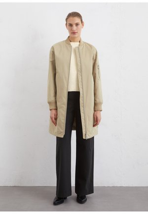 Beige bomberjack met een ritssluiting, geribde boorden en zijzakken, gedragen over een crèmekleurige trui en zwarte wijde broek.