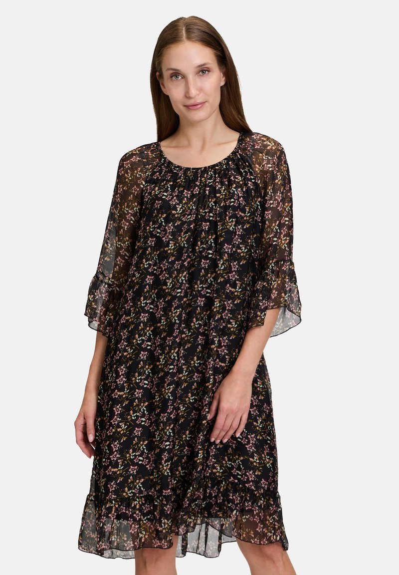 Musta kukkakuvioinen mekko, jossa on väljä istuvuus, läpikuultava kangas, poimutettu pääntie ja kellohihat. Mekossa on kukkakuosi vaaleanpunaisissa ja vihreissä sävyissä.