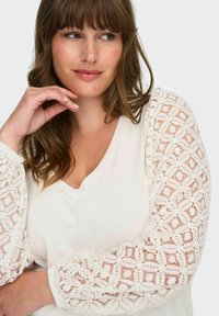 Hvit topp med gjennomsiktige blondepuffermed intrikate mønsterdetaljer, V-halsdesign og myk tekstur. Hendene hvilende under haken.