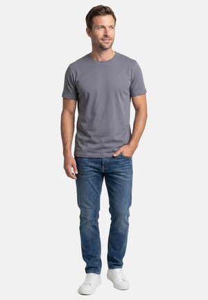 Man standing wearing a grey short-sleeve t-shirt, blue jeans, and white sneakers, with one hand in pocket, looking right.