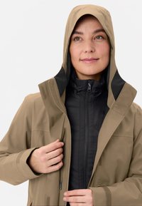 Giacca impermeabile marrone chiaro con interno nero, due tasche con zip e cappuccio. Tessuto testurizzato con cerniere visibili e dettagli in velcro.