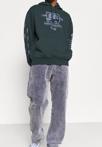 Tmavě zelená mikina s kapucí s grafickým potiskem "DIGITAL DEVIANCE" a texturovanými akcenty. Kombinováno s světle šedými manšestrovými kalhotami.