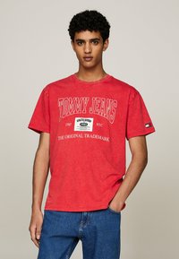 Camiseta de algodón roja con mangas cortas, que presenta el texto blanco "TOMMY JEANS" y un gráfico de logo. Combinada con unos vaqueros de denim azul.